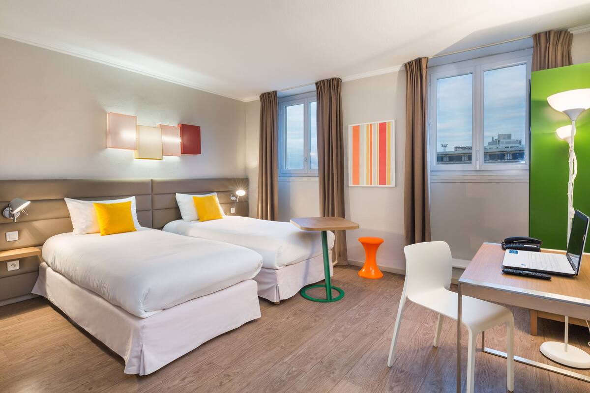 A hotel room with two beds, a desk with a laptop, a chair, a small table, and a lamp.