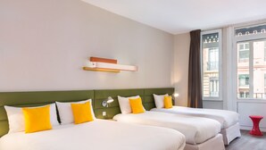 A hotel room with two beds, a green headboard, and a window with a view of buildings.