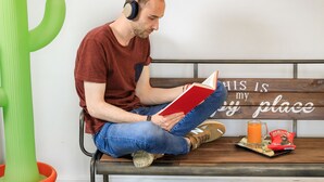 A man sitting on a bench reading a book, with a foosball table and a cactus in the room.