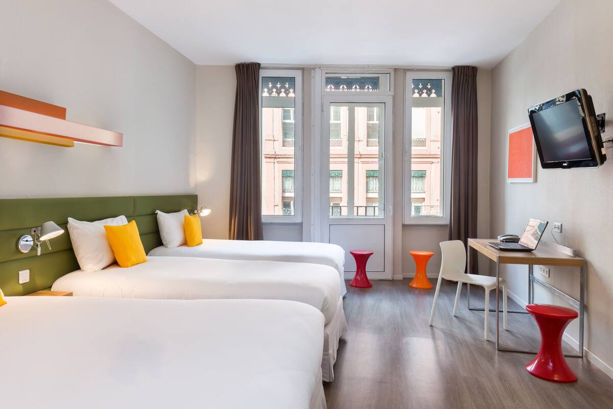A hotel room with two beds, a desk, a chair, a TV, and a window with curtains.