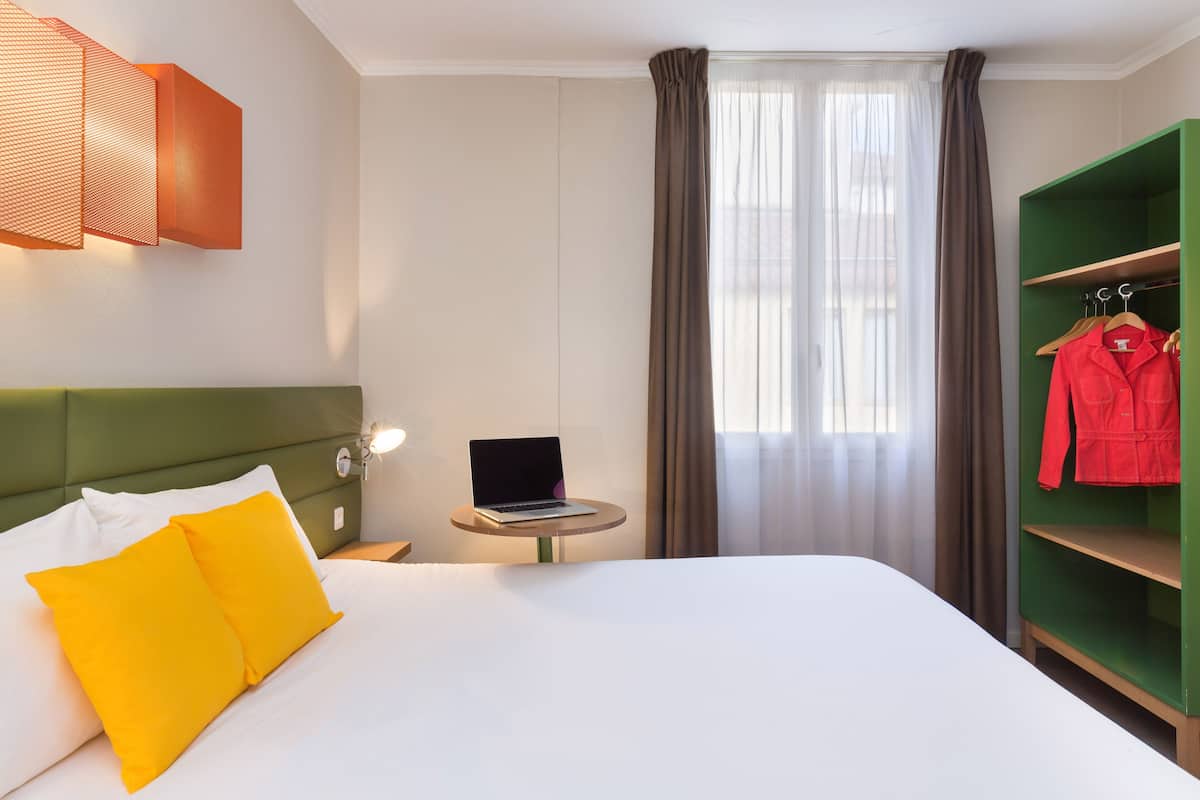 A hotel room with a bed, a laptop on a small table, a green wardrobe, and a red jacket hanging on the wardrobe.