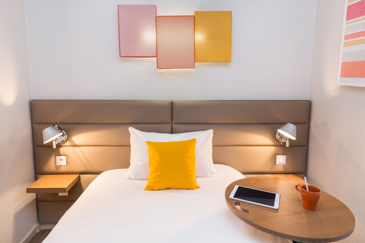 A modern hotel room with a bed, bedside tables, a bedside lamp, a tablet, and a cup.