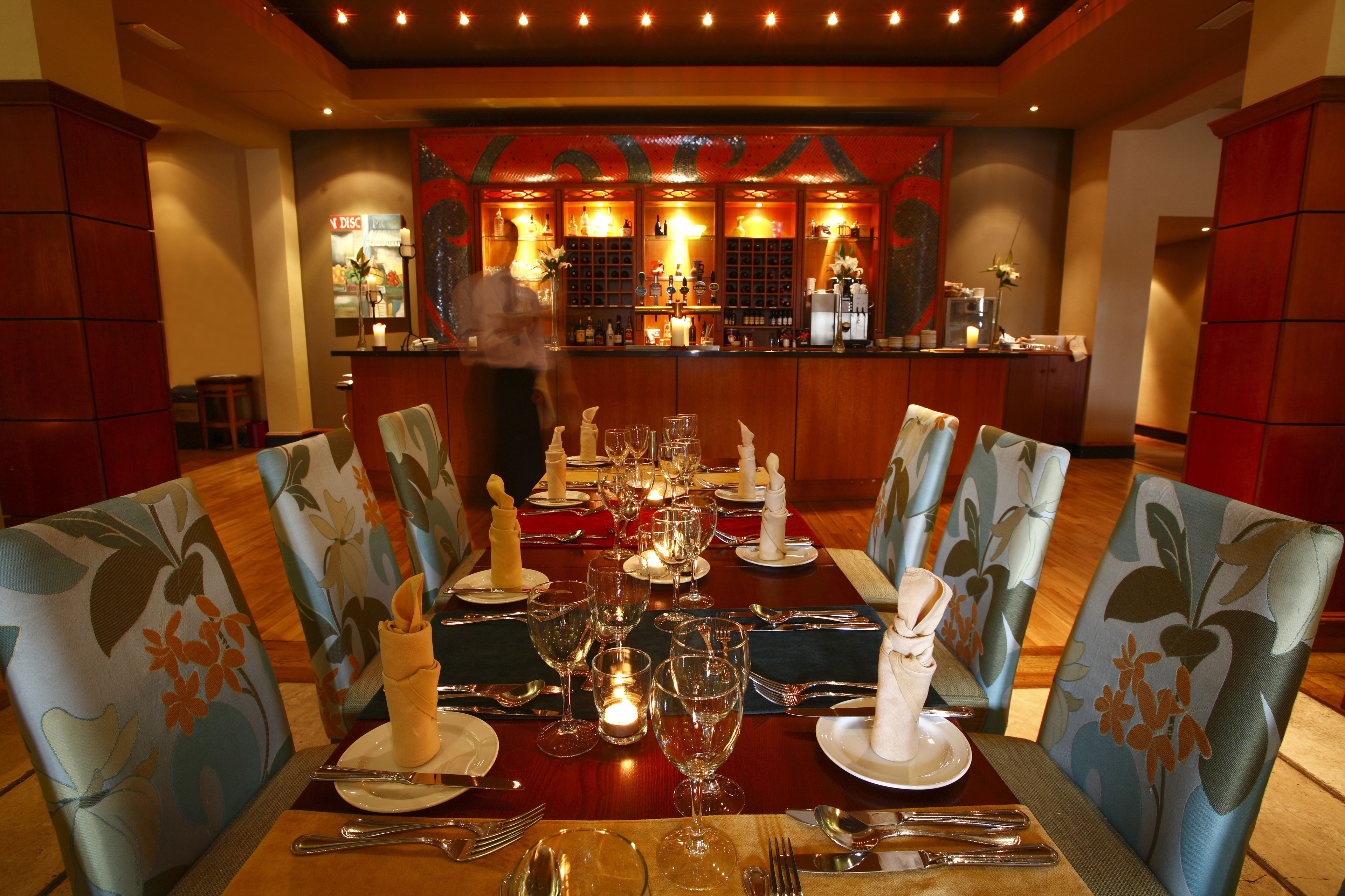 A restaurant with a long table set for a meal, chairs with floral upholstery, and a bar in the background.