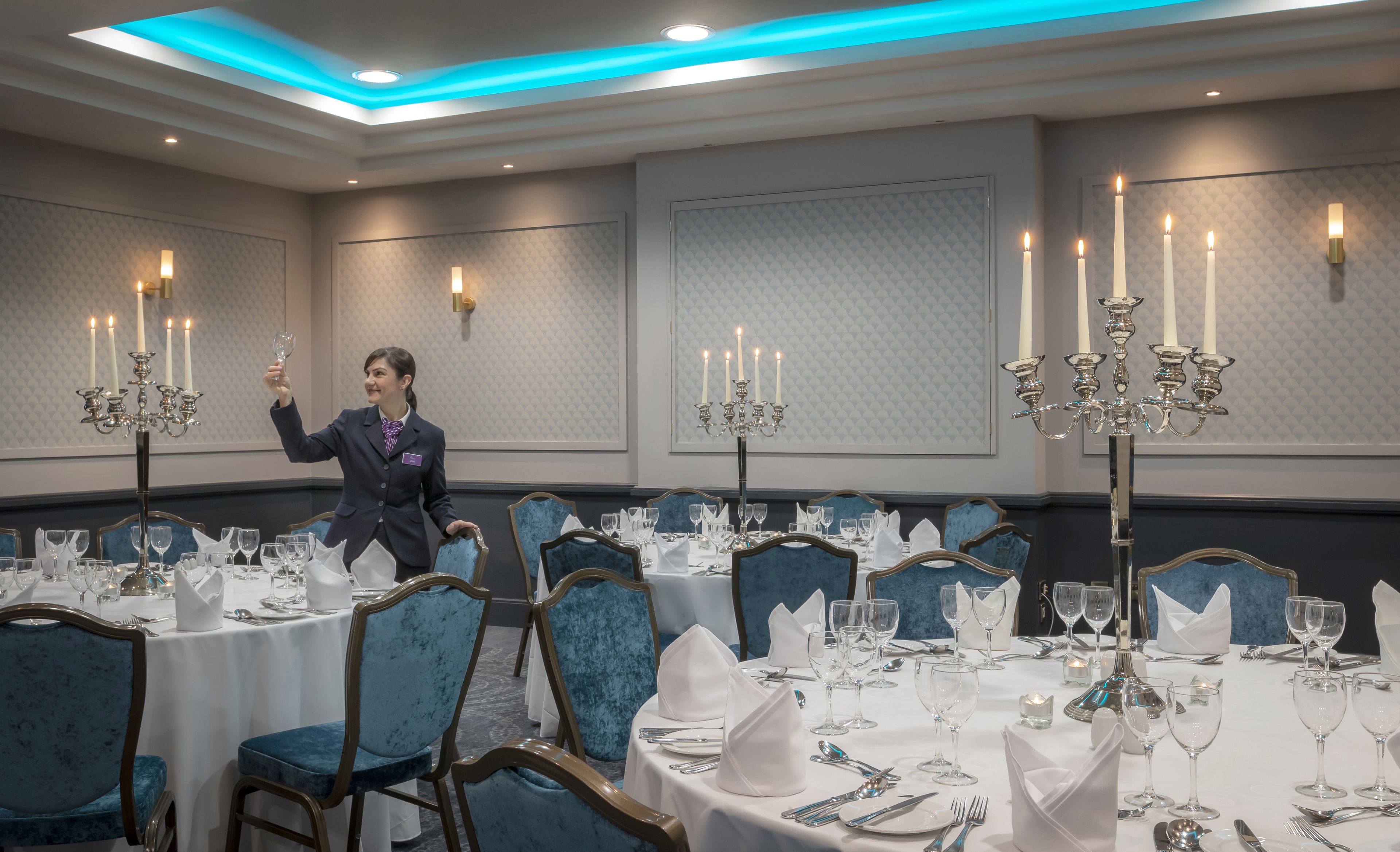 A banquet hall with round tables set for dining, blue upholstered chairs, and chandeliers with multiple candles.