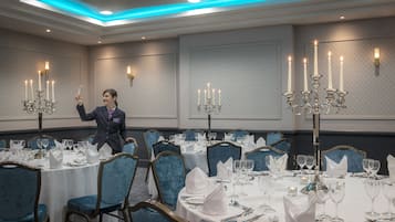 Un salón de banquetes con mesas redondas dispuestas para comer, sillas tapizadas en azul y candelabros con múltiples velas.