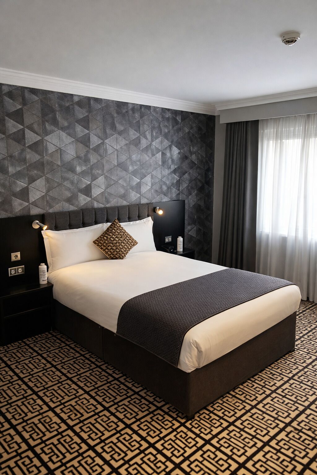 A hotel room with a large bed, a geometric-patterned wall, a nightstand with a lamp, and a patterned floor.