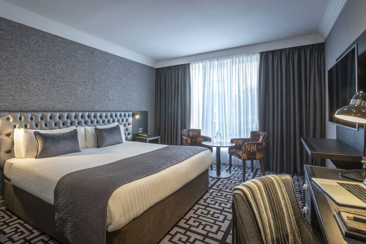 A hotel room with a large bed, a desk, a chair, and a small table.