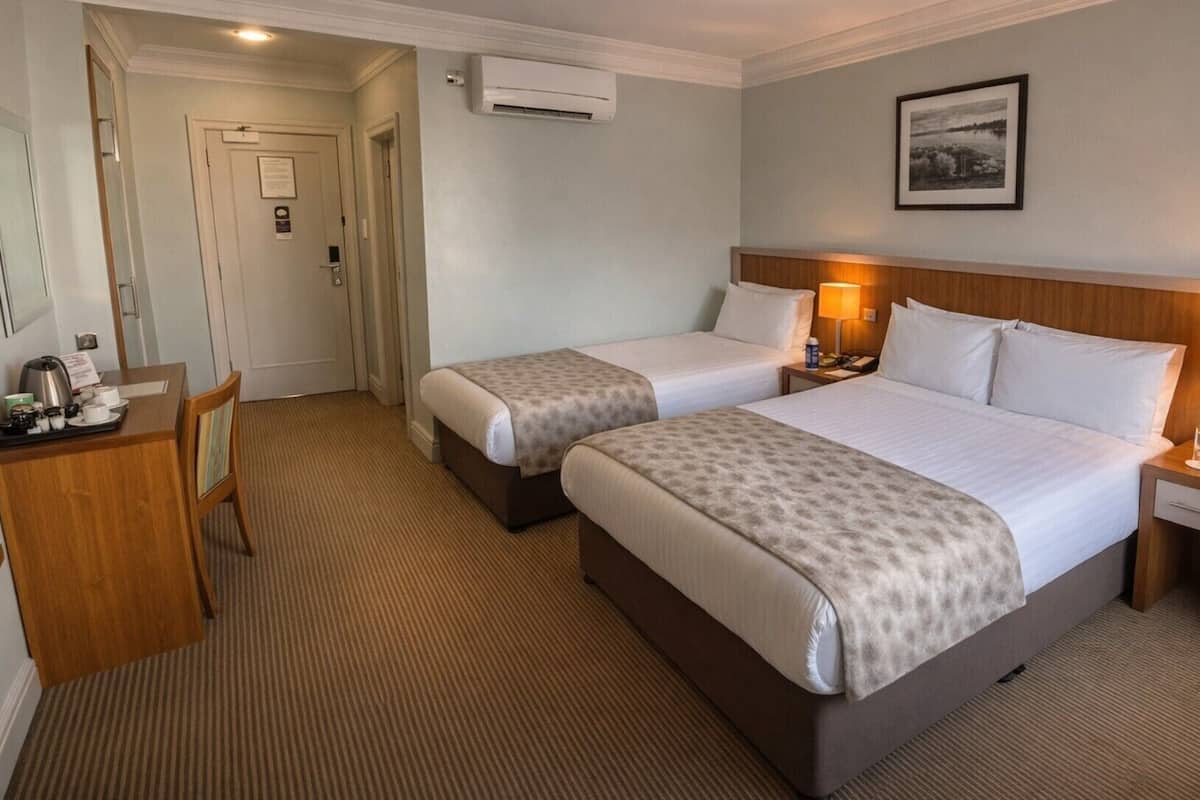 A hotel room with two beds, a desk with a chair, a bedside table with a lamp, and a framed picture on the wall.