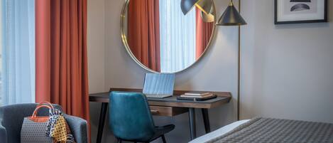 Een hotelkamer met een bed, een bureau met een laptop, een stoel, een ronde spiegel, een lamp en een ingelijst schilderij aan de muur.