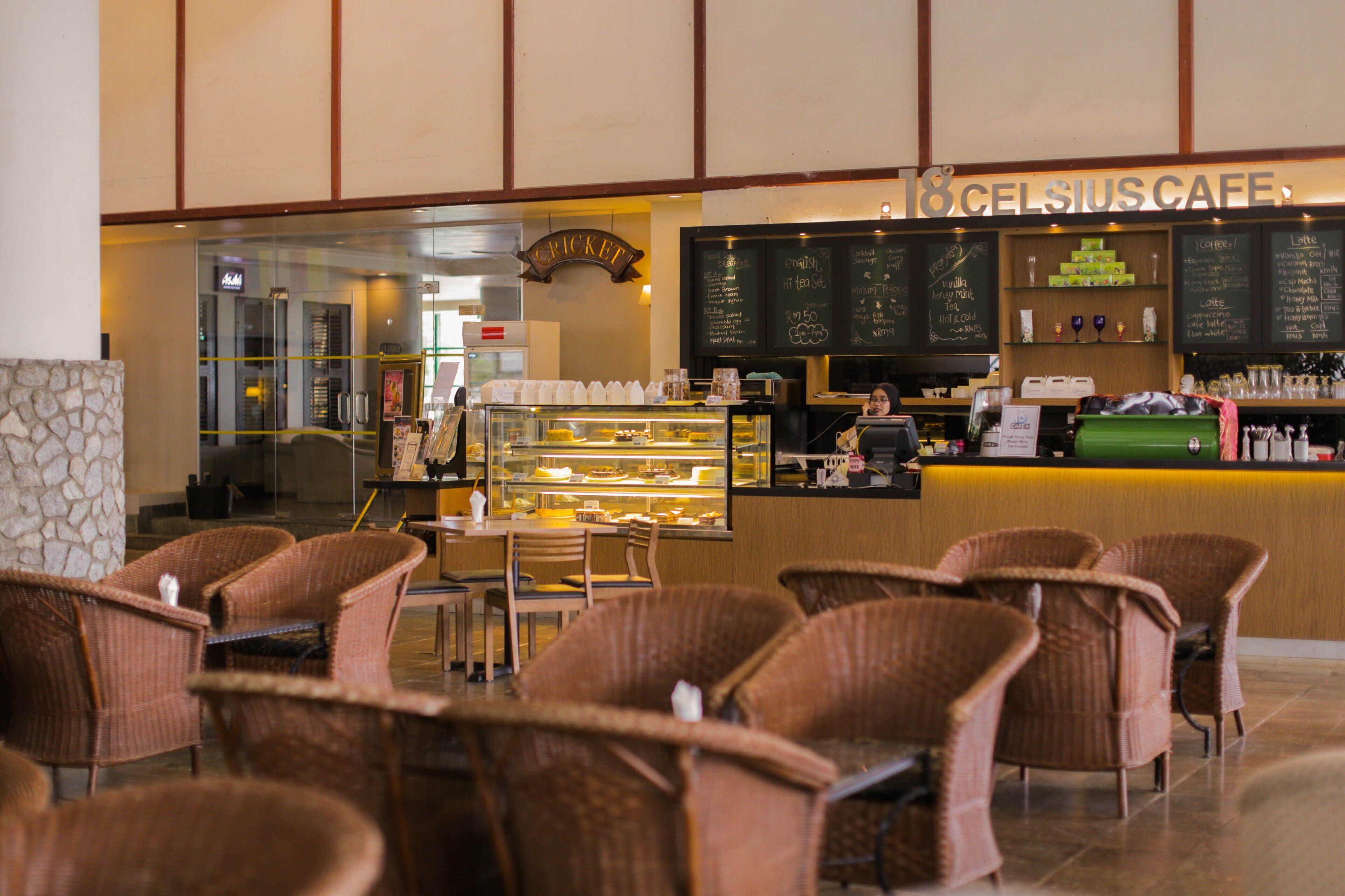 Una cafetería con sillas de mimbre, una barra con menú en pizarra y una vitrina con productos alimenticios.