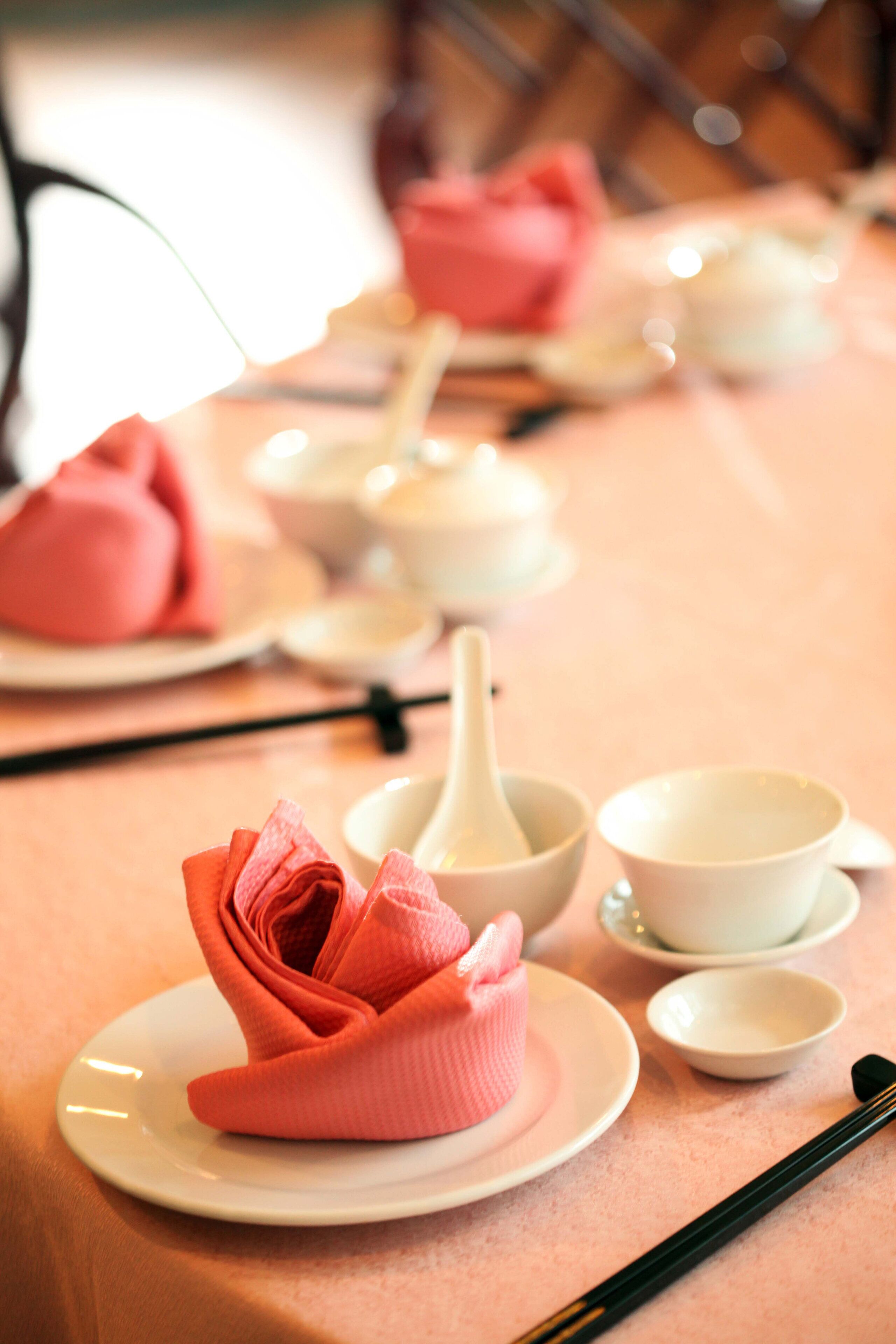 Una mesa con mantel rosa, un plato con un objeto rosa doblado y varios cuencos blancos y una cuchara.