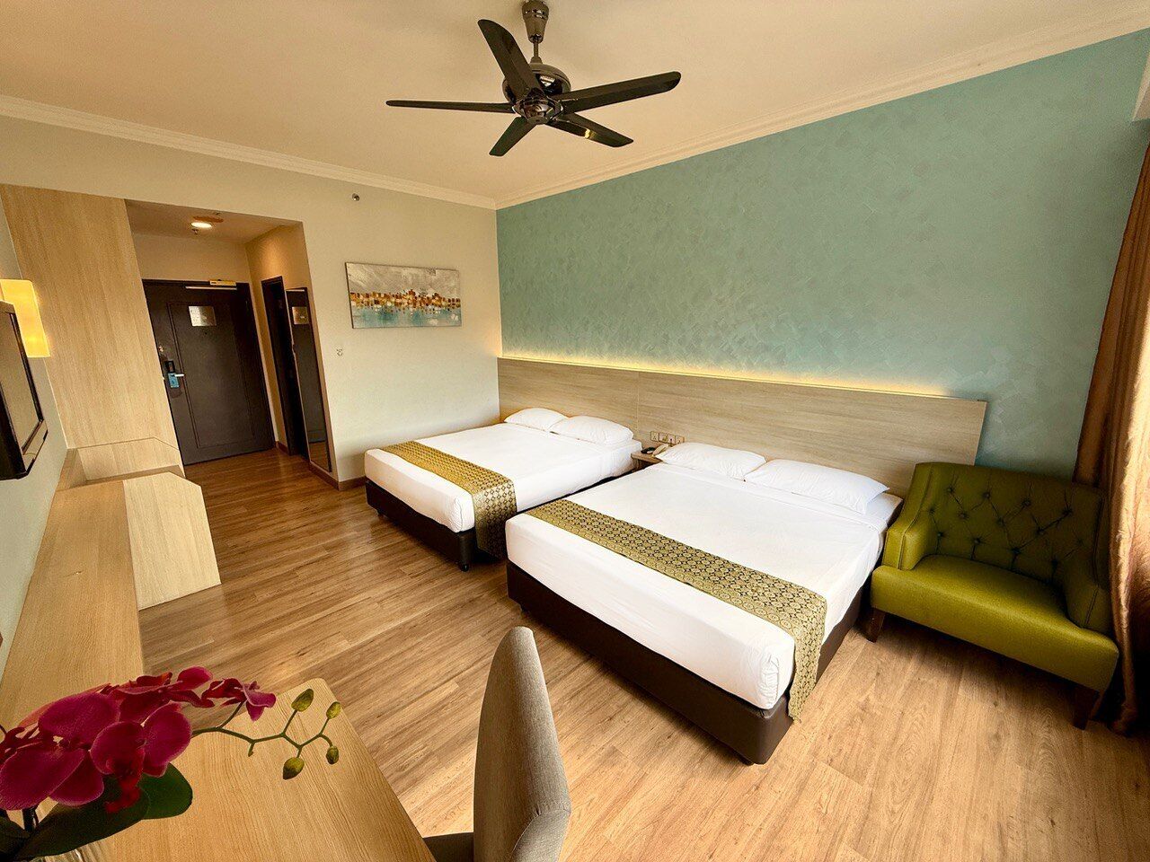 Ein Hotelzimmer mit zwei Betten, einem grünen Sessel, Holzboden, Deckenventilator und einem Gemälde an der Wand.