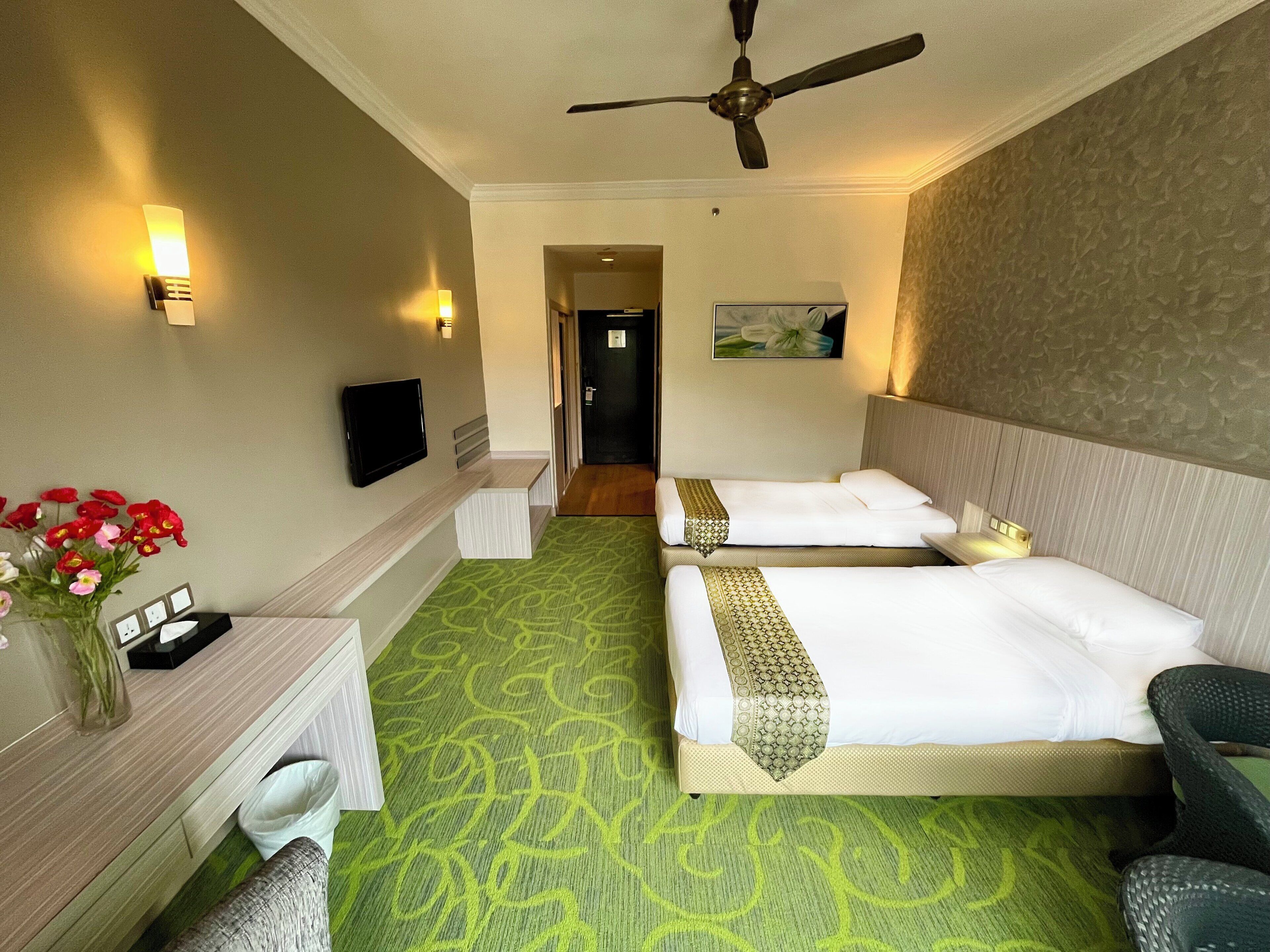 A hotel room with two beds, a desk, a chair, a TV, and a ceiling fan.
