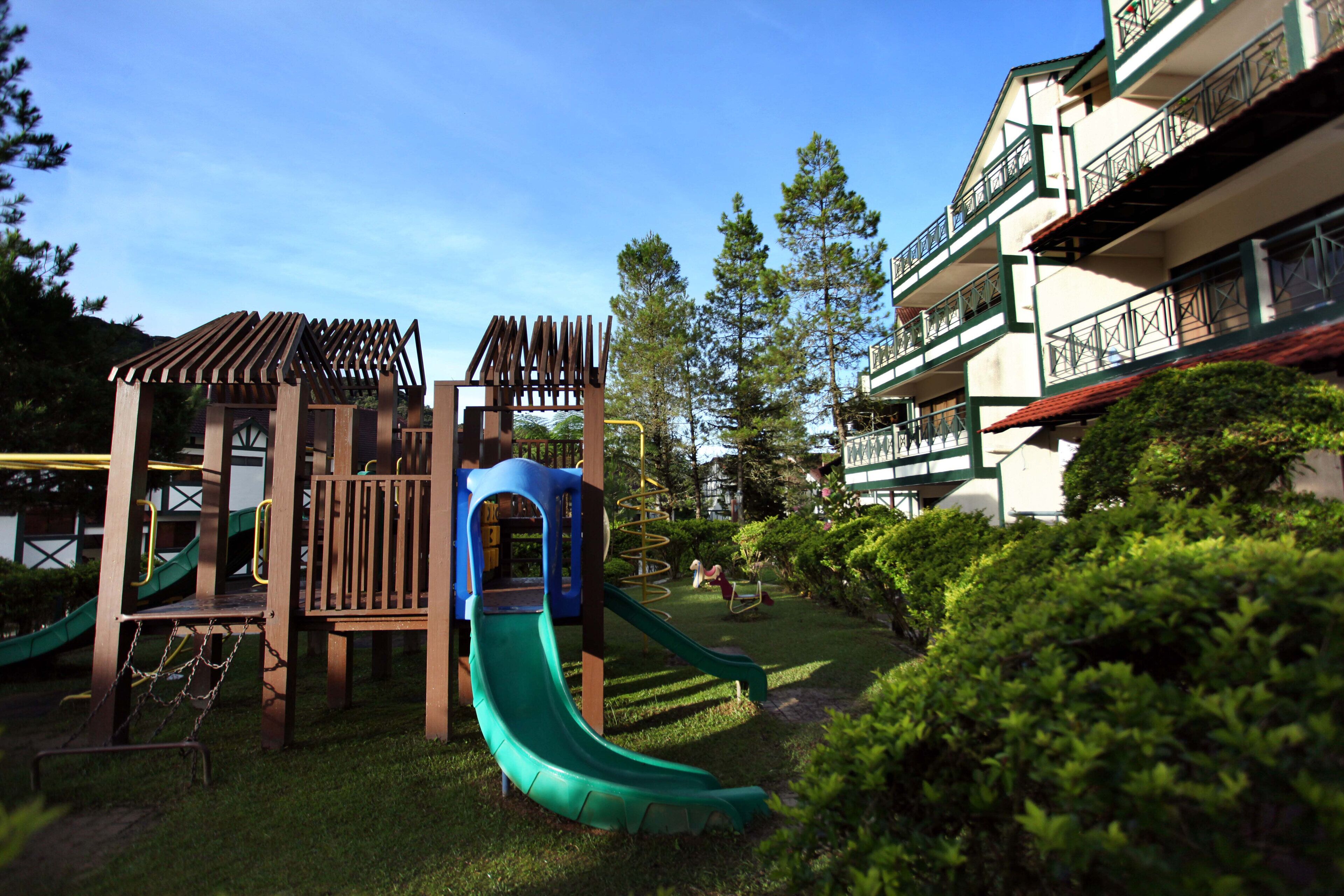 Un parque infantil con toboganes y estructuras para escalar, rodeado de vegetación y un edificio con balcones.