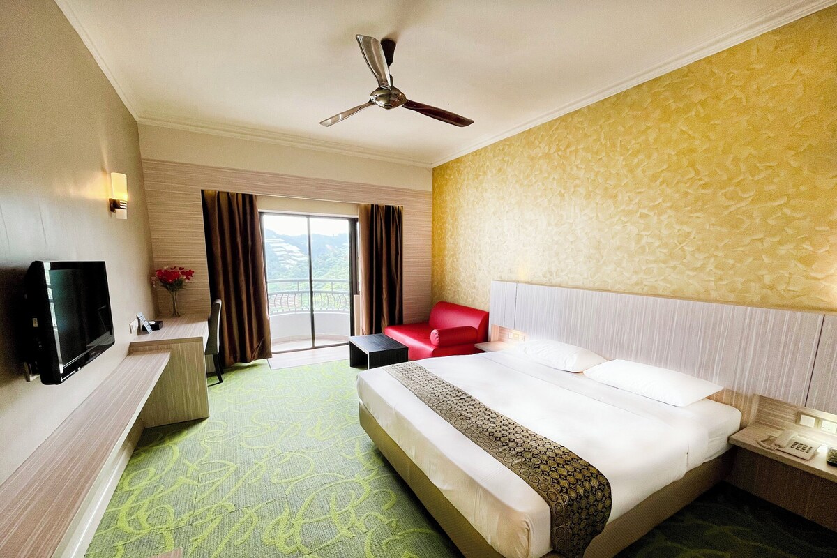 Habitación de hotel con cama, escritorio, televisor, una silla roja y balcón con cortinas.