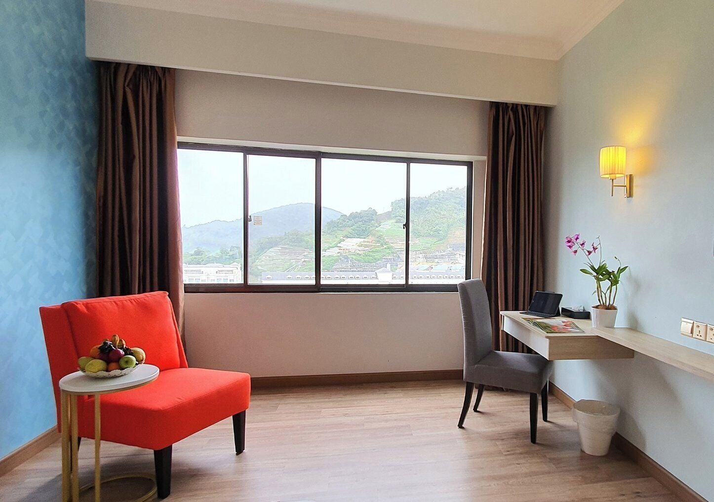 Une chambre d’hôtel avec une grande fenêtre, un fauteuil rouge, un fauteuil gris, une petite table sur laquelle se trouve un panier de fruits, un bureau avec un ordinateur portable et une plante en pot.