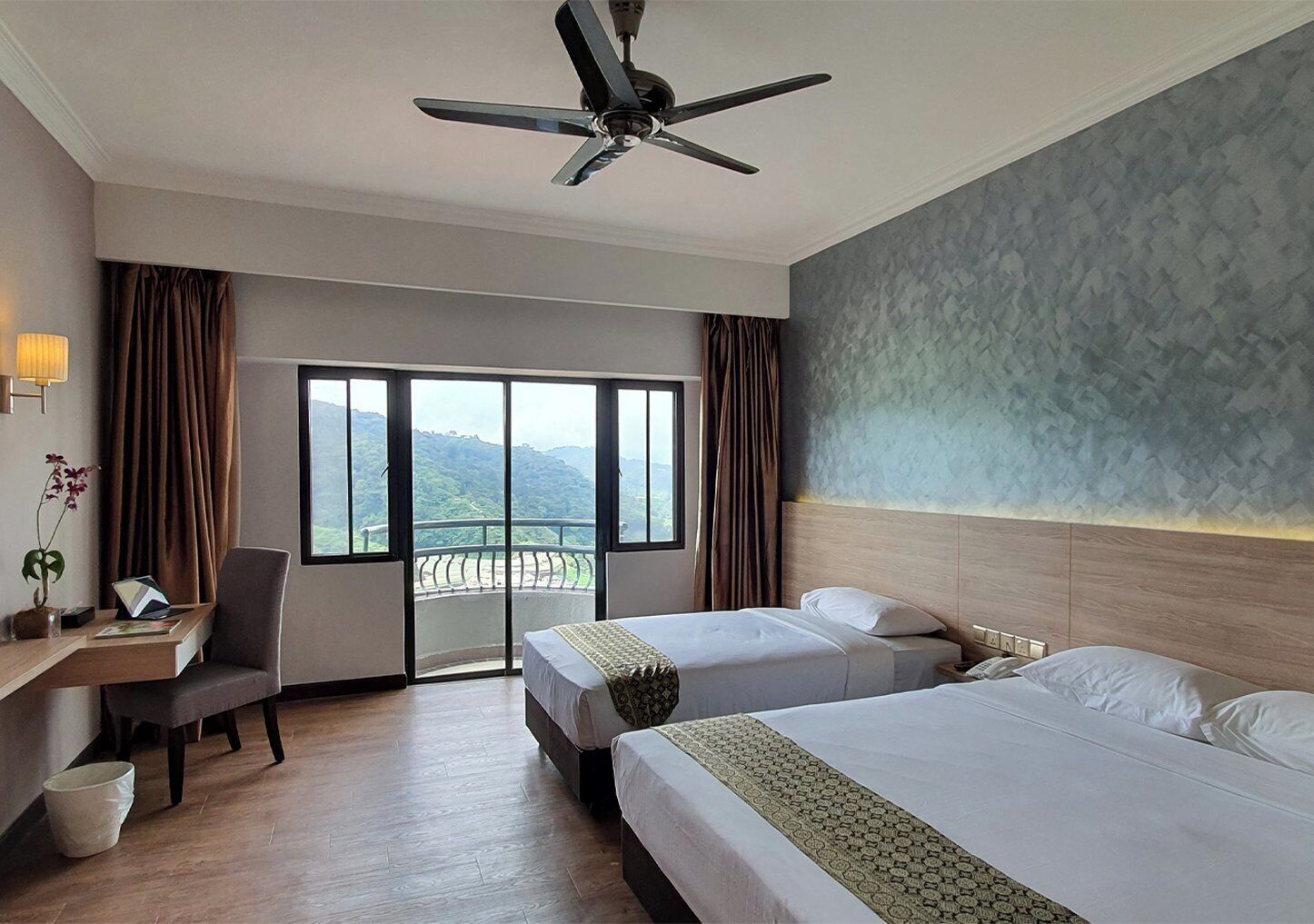 A hotel room with two beds, a desk, a chair, a ceiling fan, and a large window with a view of the mountains.