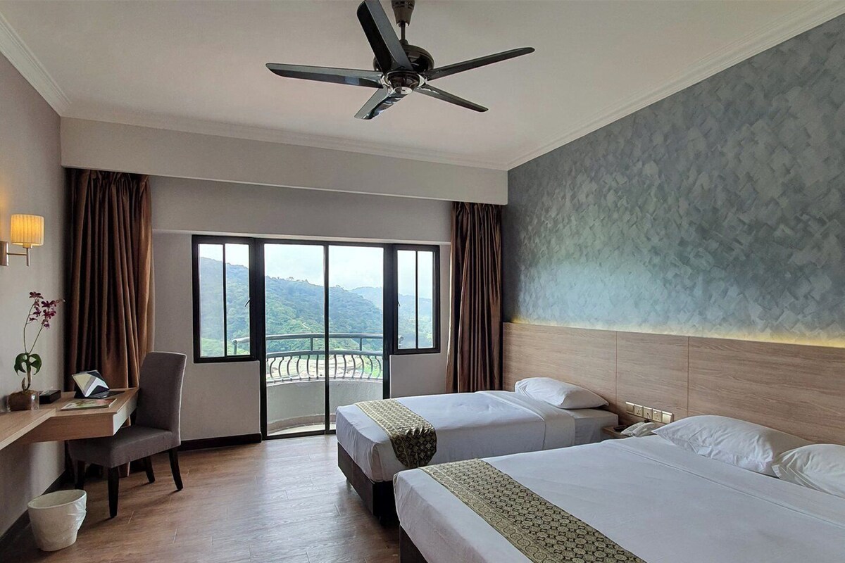Habitación de hotel con dos camas, un escritorio, una silla, un ventilador de techo y un ventanal con vistas a las montañas.