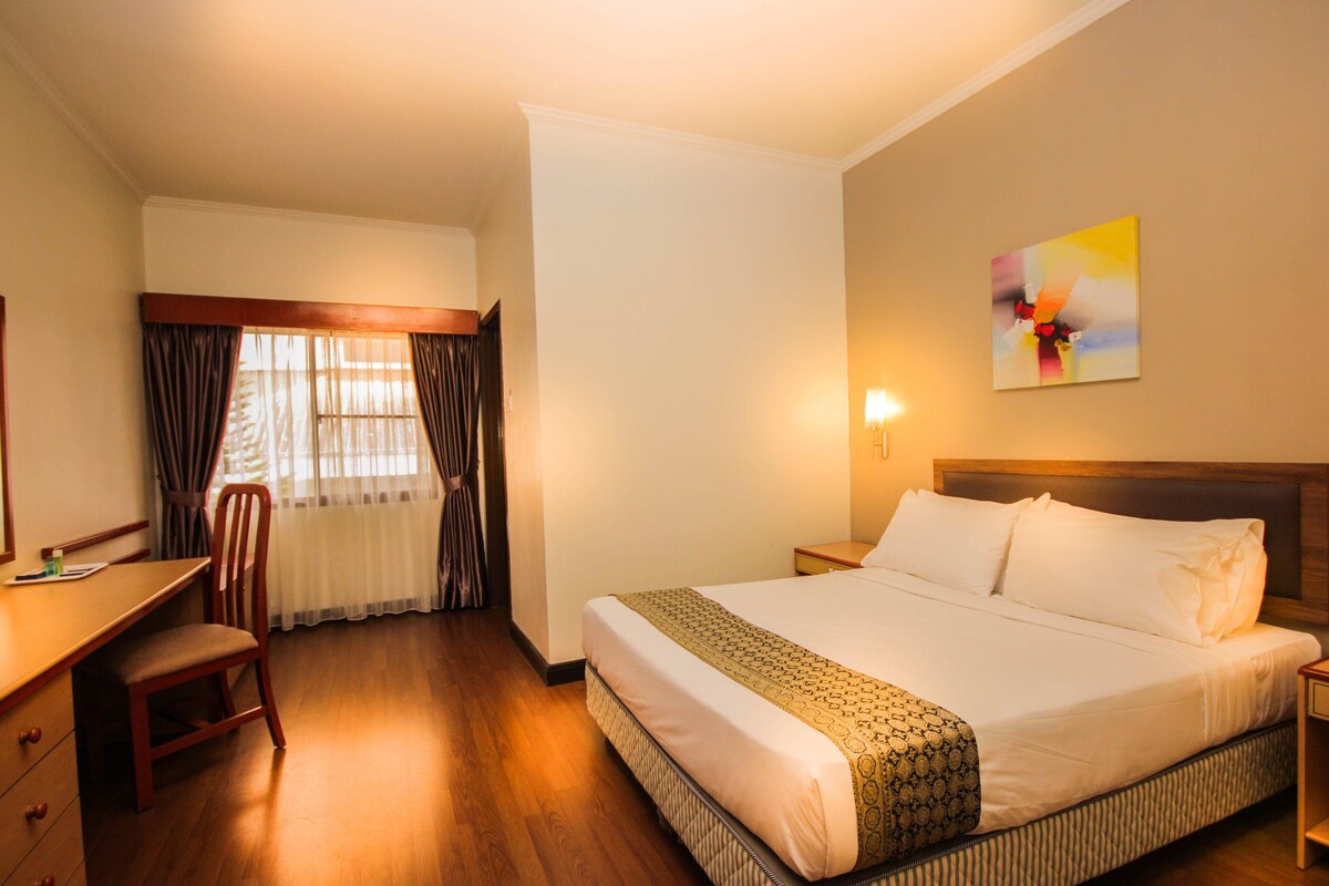 Una habitación de hotel con cama, escritorio, silla, ventana con cortinas y un cuadro en la pared.
