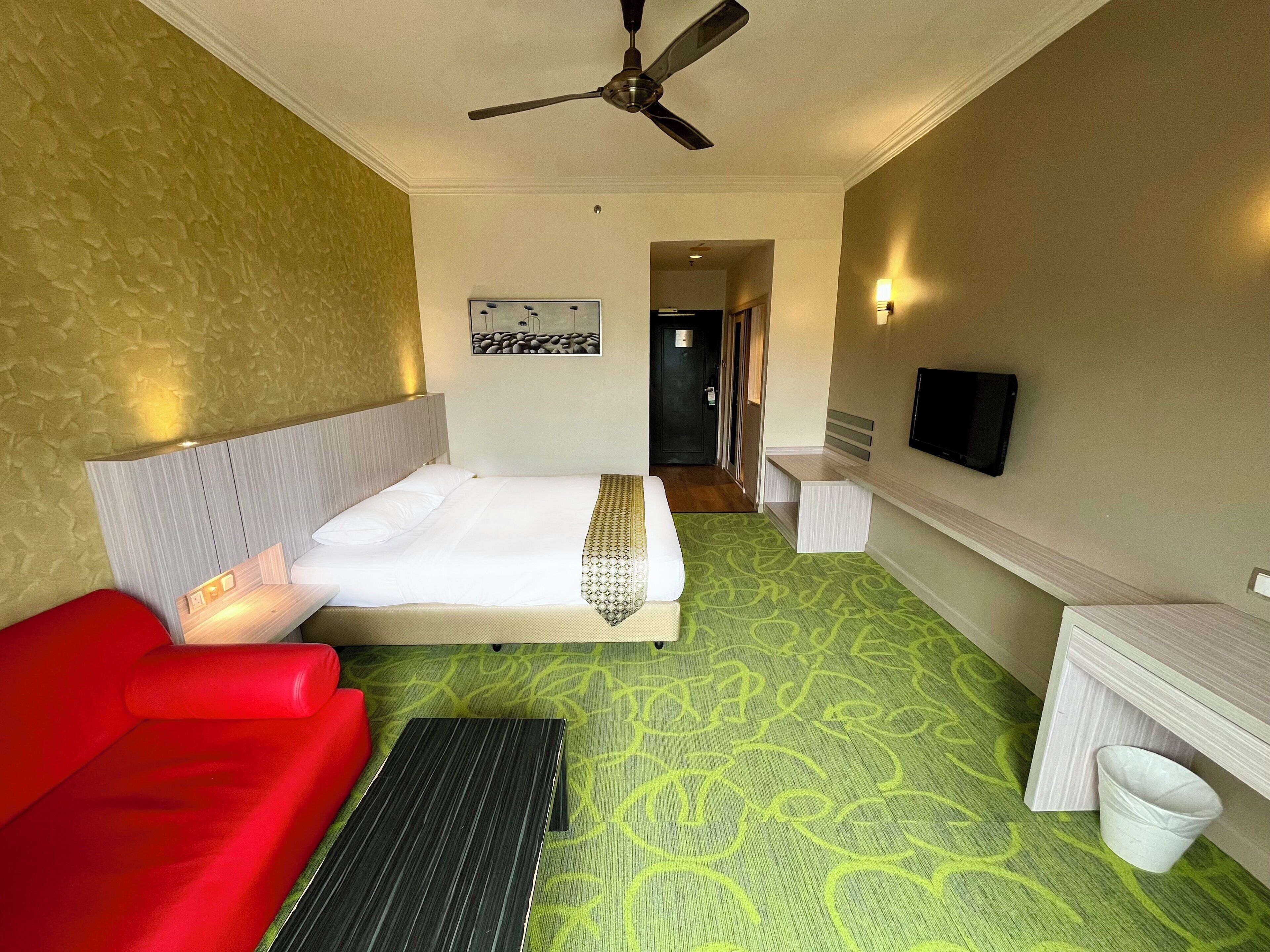 A hotel room with a bed, a red sofa, a desk, a ceiling fan, and a television.