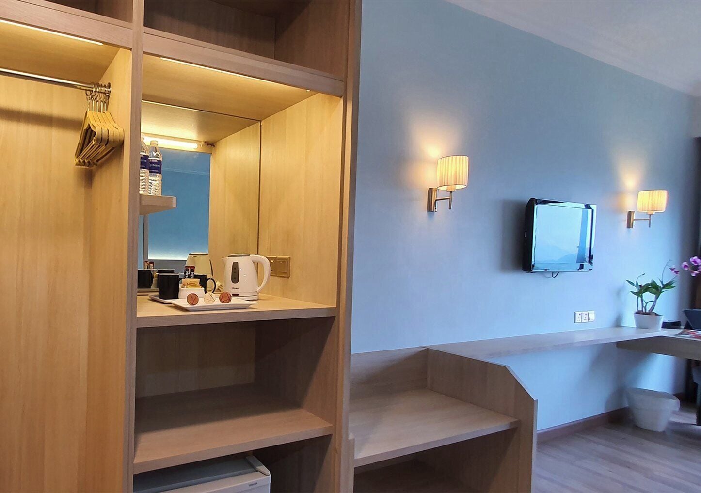 A hotel room with a wooden wardrobe, a mirror, a television mounted on the wall, and a small table with a kettle and cups.