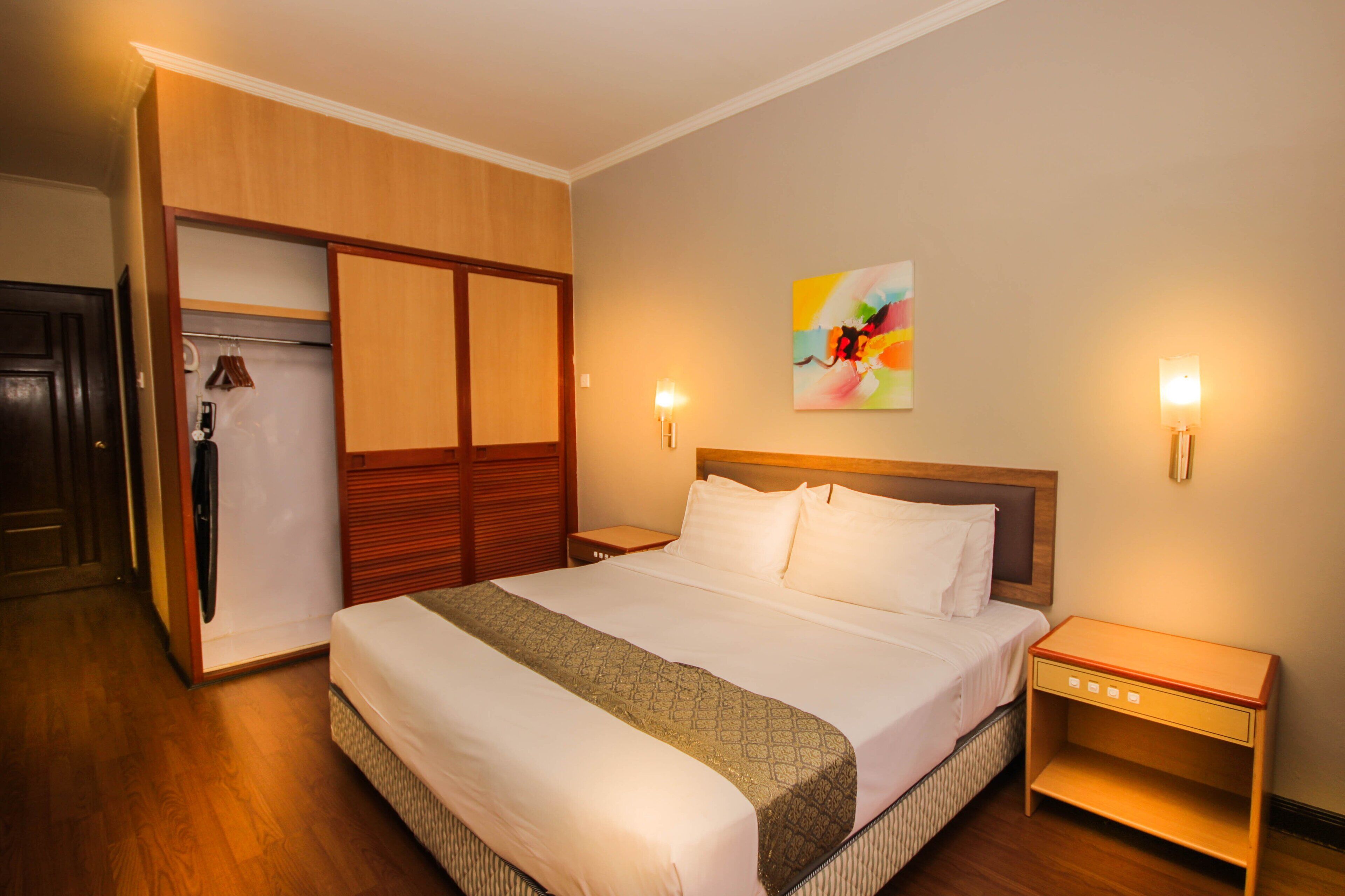 A hotel room with a bed, wooden wardrobe, bedside tables, and a colorful abstract painting on the wall.