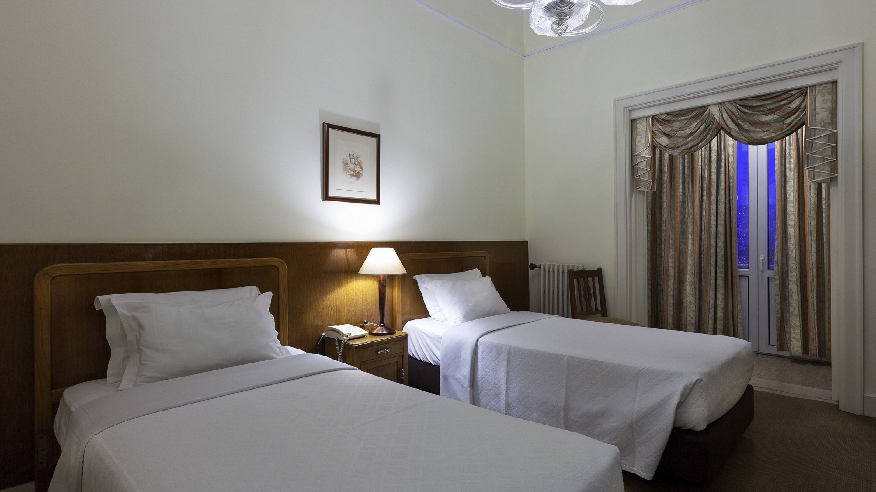 Une chambre d’hôtel avec deux lits simples, une table de chevet avec une lampe, un tableau encadré au mur et une fenêtre avec des rideaux.