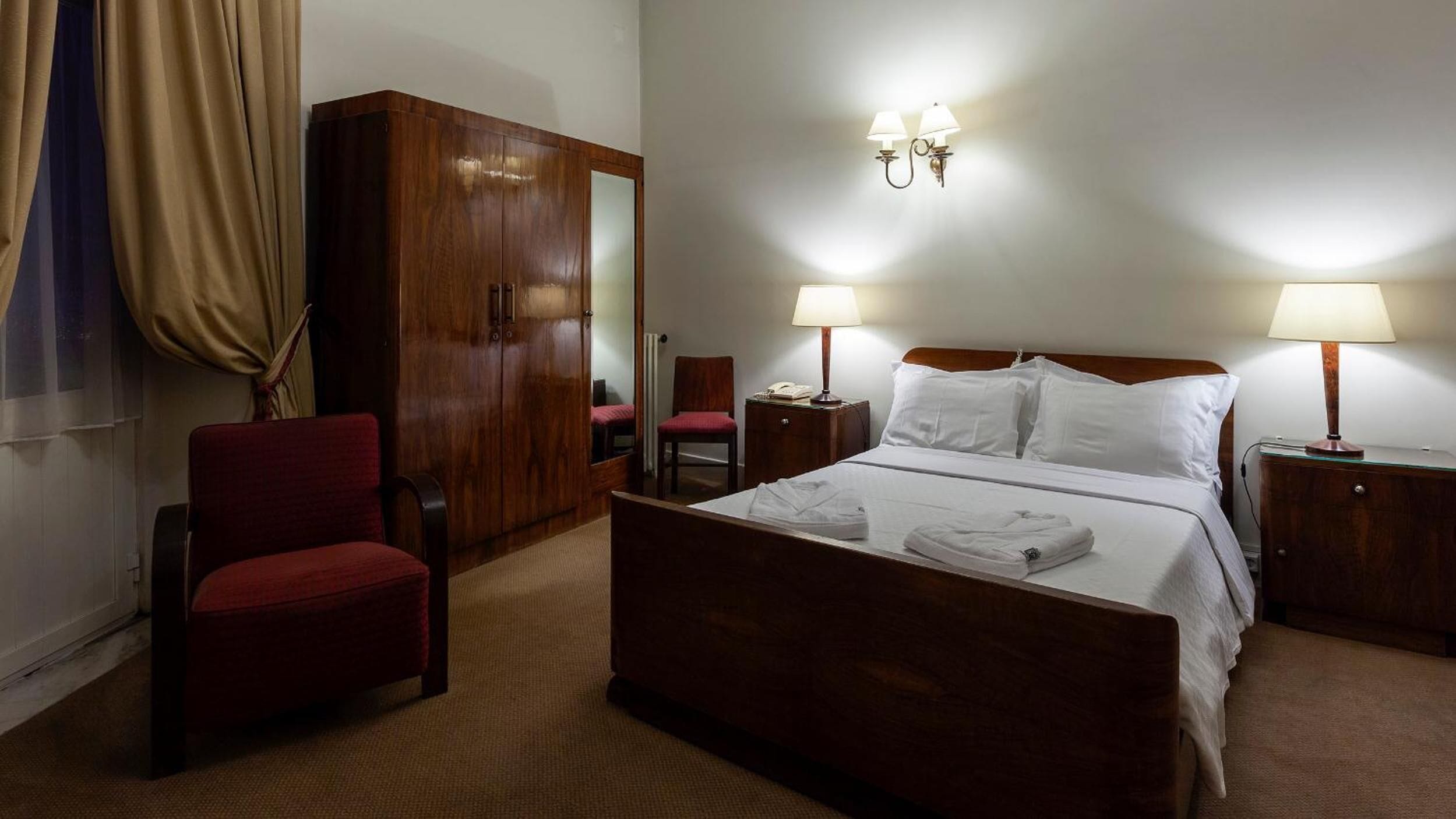 A hotel room with a bed, two bedside tables, a wardrobe, a chair, and two wall-mounted lamps.
