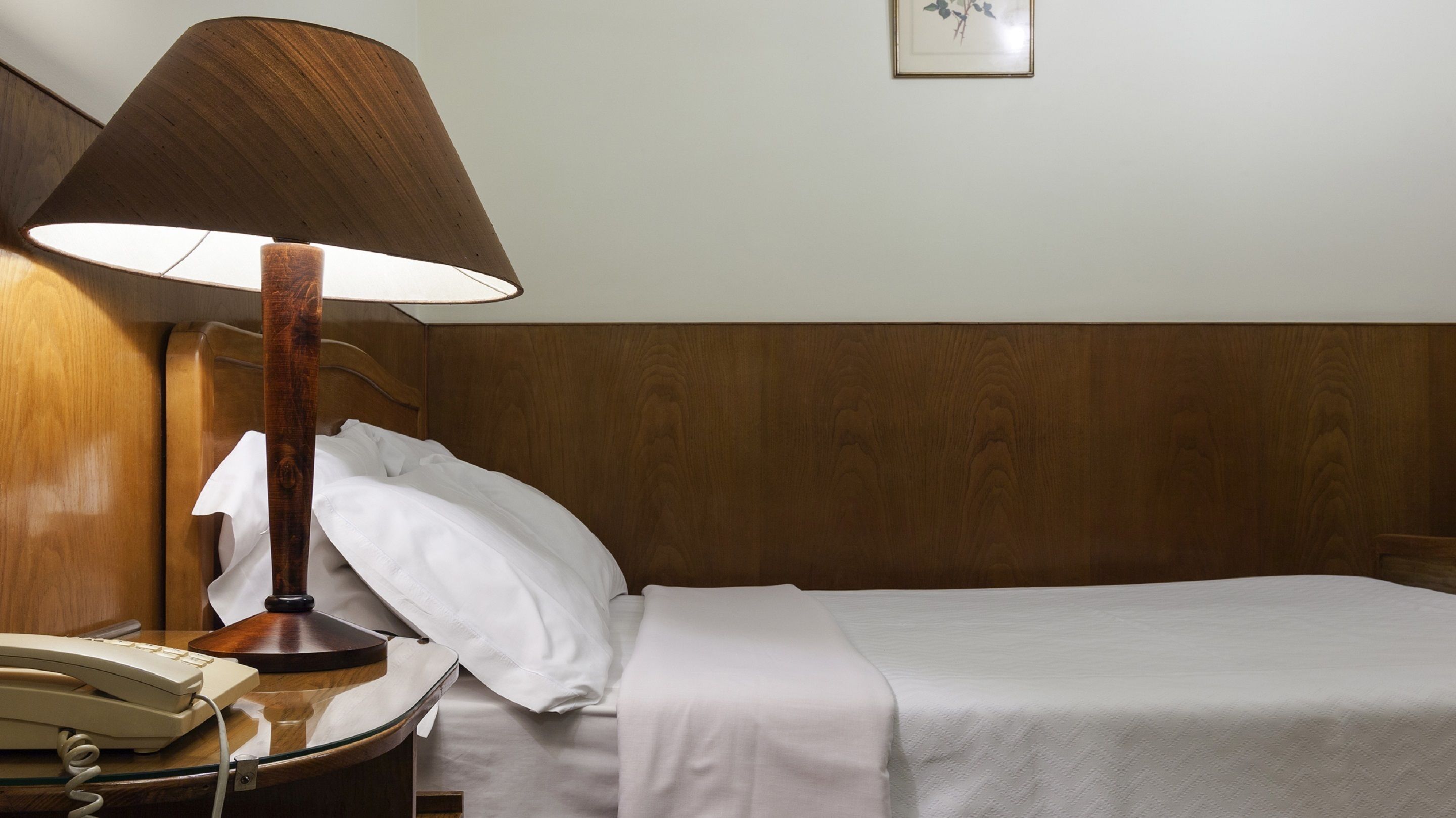 A neatly made bed with white linens, a wooden headboard, a bedside lamp, and a telephone on a wooden nightstand.