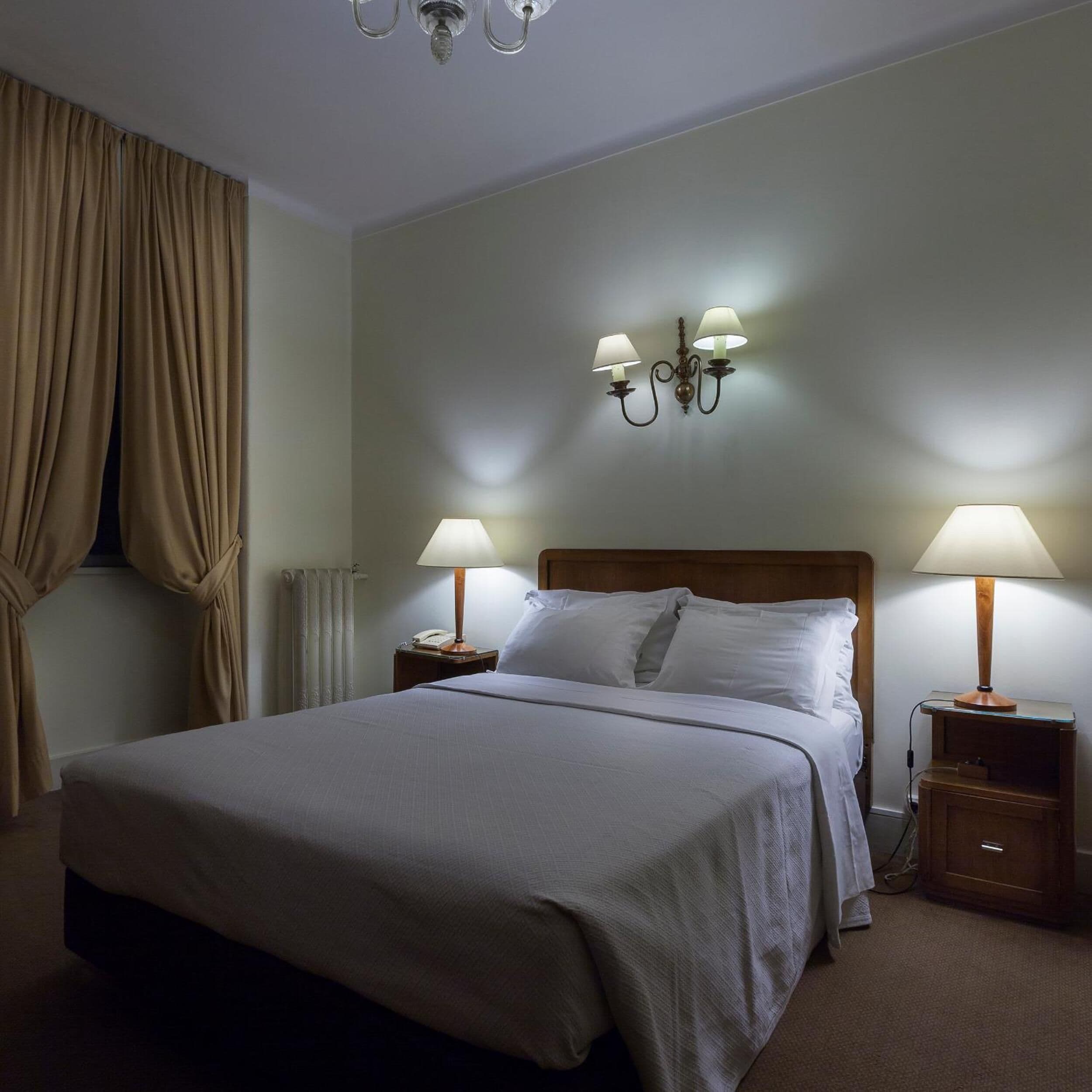 A hotel room with a large bed, two bedside tables with lamps, a wall-mounted light fixture, and a window with curtains.