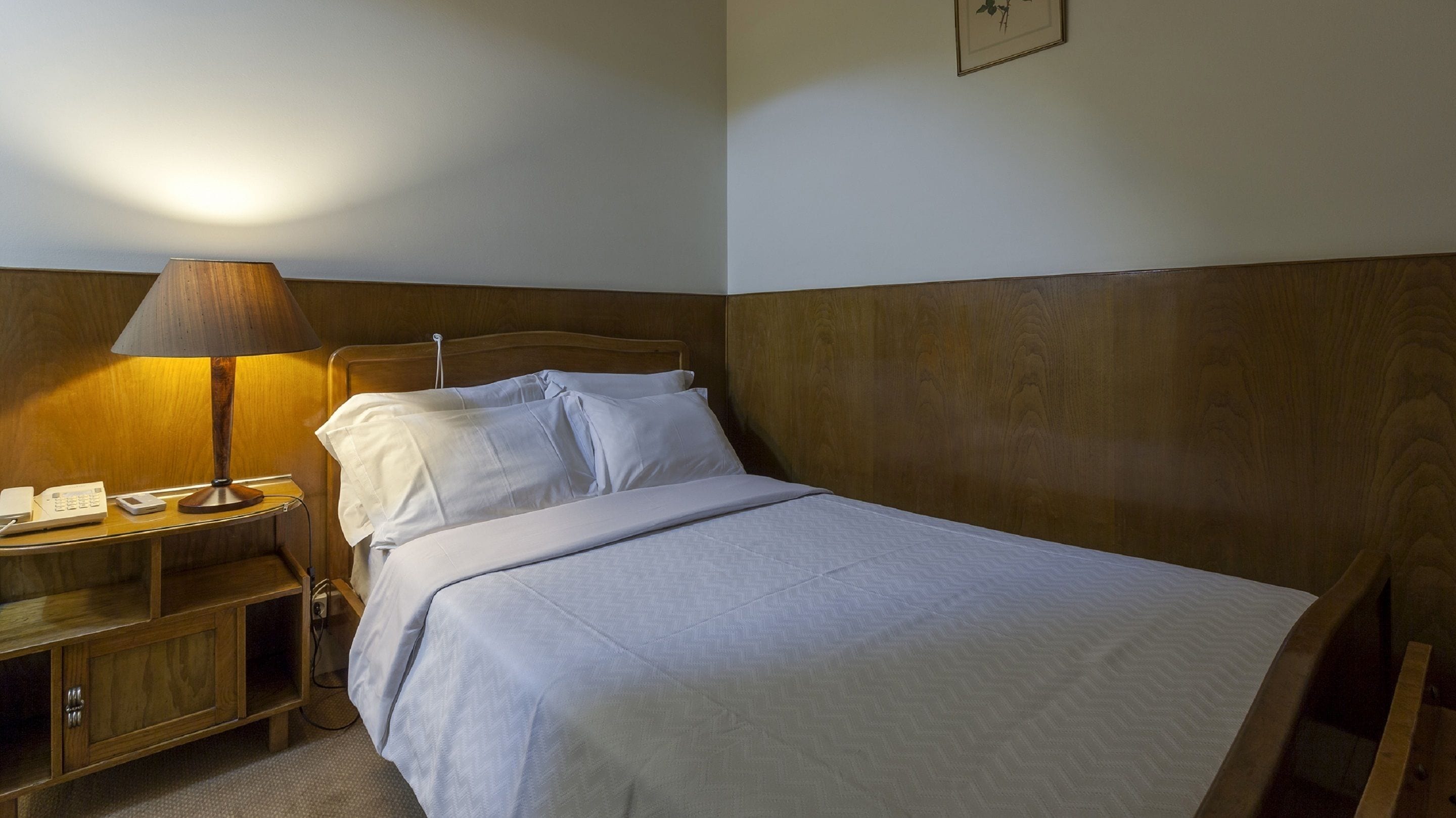 Una cama bien hecha con ropa de cama blanca, una mesita de noche de madera con una lámpara y un teléfono.