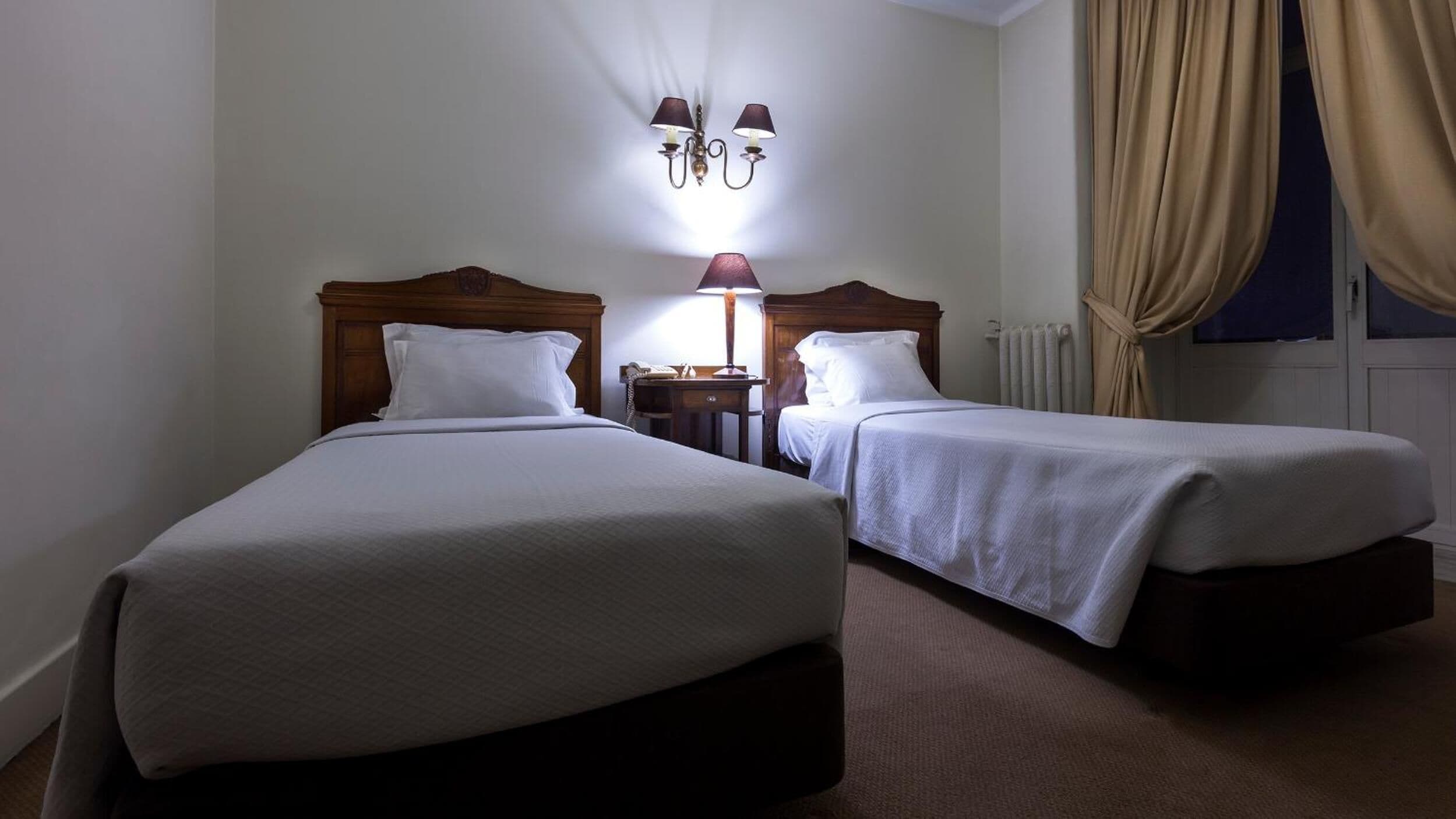 Habitación de hotel con dos camas, una mesita de noche con lámpara, un radiador y una ventana con cortinas.