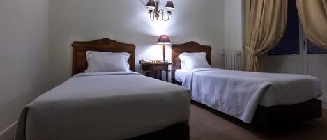 Habitación de hotel con dos camas, una mesita de noche con lámpara, un radiador y una ventana con cortinas.