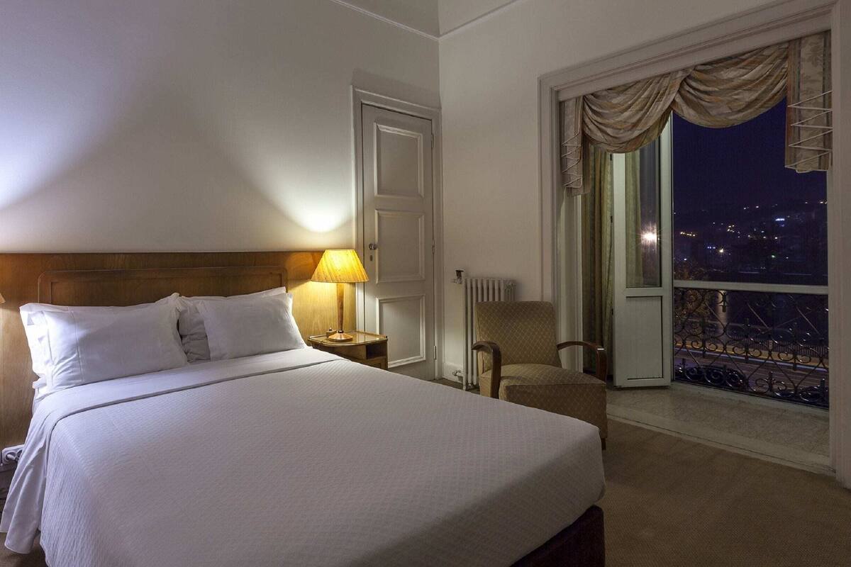 A hotel room with a bed, two bedside lamps, a chair, a nightstand, a radiator, and a window with a view of the city at night.