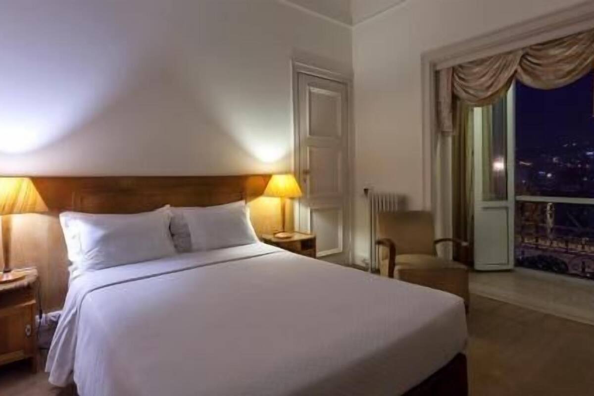 A hotel room with a bed, two bedside lamps, a nightstand, a chair, and a window with a view.