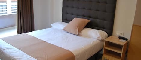 A neatly made bed with a headboard, a bedside table with a phone, and a window with curtains.