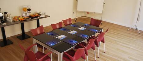 A conference room with a long table, red chairs, a whiteboard, and a buffet table with fruits and beverages.