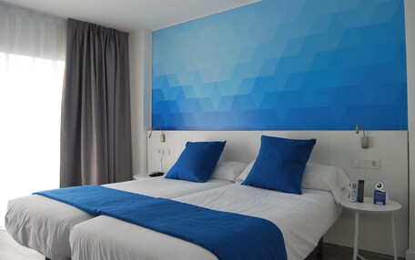 A hotel room with two beds, a large blue geometric wall art, a white bedside table with a remote control, and a grey curtain.