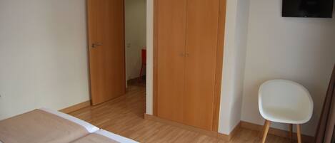 A bedroom with a bed, a wooden wardrobe, a white chair, and a television on the wall.
