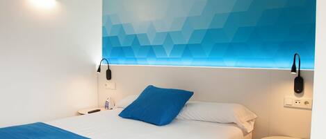 Habitación de hotel con cama, almohada azul, mesita de noche y lámpara fijada a la pared.