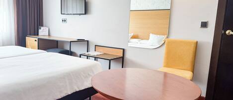 A hotel room with a large bed, a desk, a chair, a round table, and a wall-mounted TV.