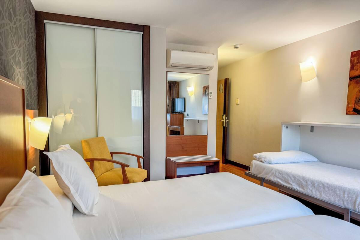 Quarto de hotel com duas camas, uma cadeira, uma escrivaninha e um espelho.
