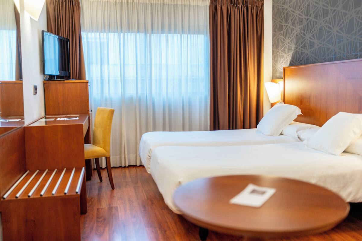 Quarto de hotel com cama de madeira, mesa de cabeceira, uma cadeira, televisão sobre um armário de madeira e janela com cortinas.
