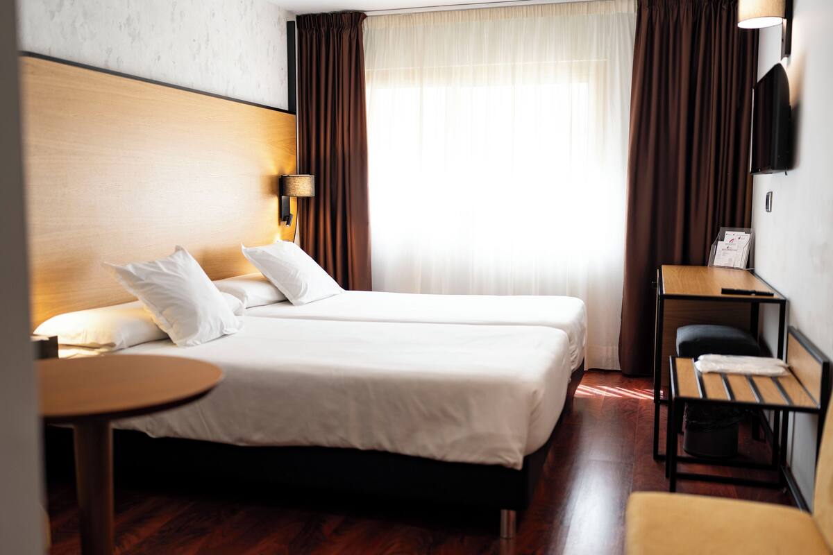 Quarto de hotel com duas camas, uma mesinha, uma cadeira, uma escrivaninha e televisão presa na parede.