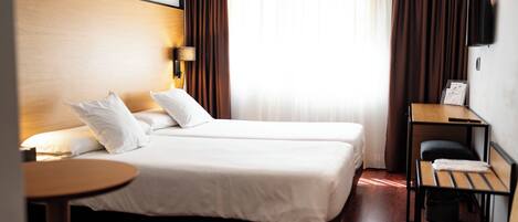 A hotel room with two beds, a small table, a chair, a desk, and a wall-mounted TV.