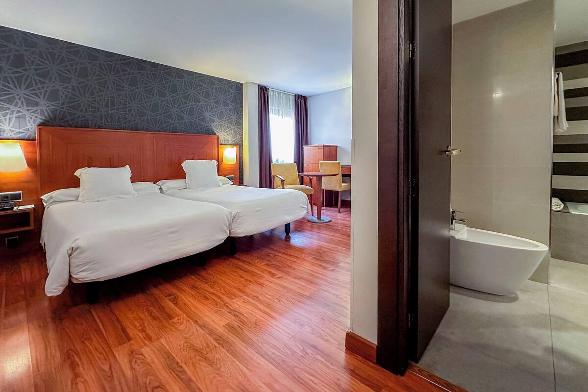 Quarto de hotel com duas camas, piso de madeira, banheiro moderno com banheira e parede estampada.