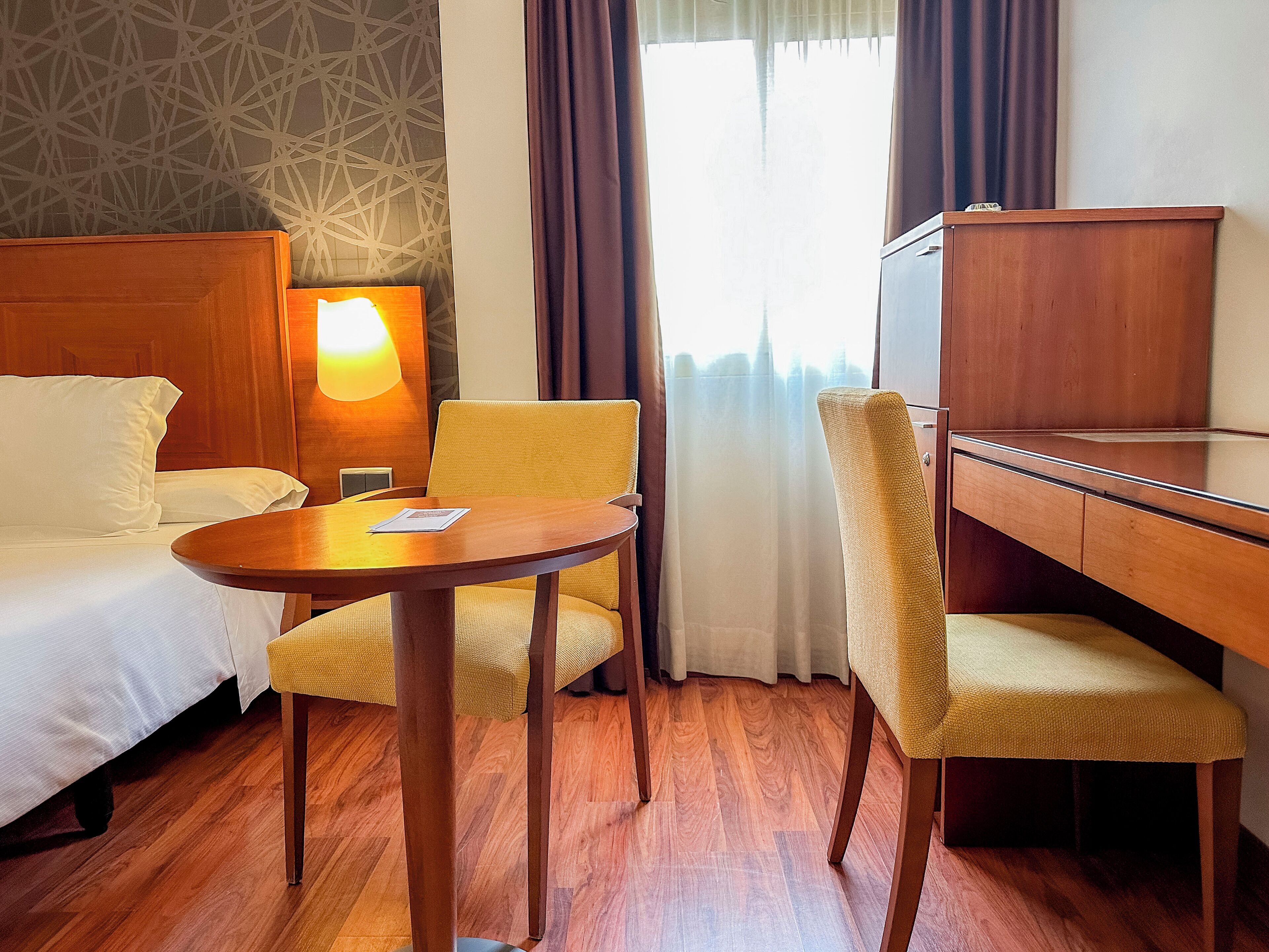 A hotel room with a bed, a desk, two chairs, a lamp, and a window with curtains.