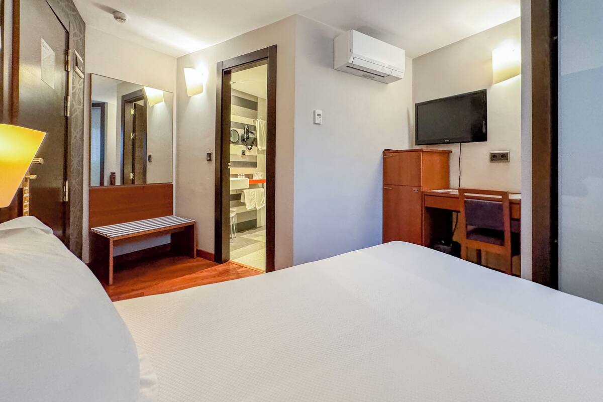 Quarto de hotel com uma cama, um banquinho, uma escrivaninha, uma televisão e um guarda-roupa.