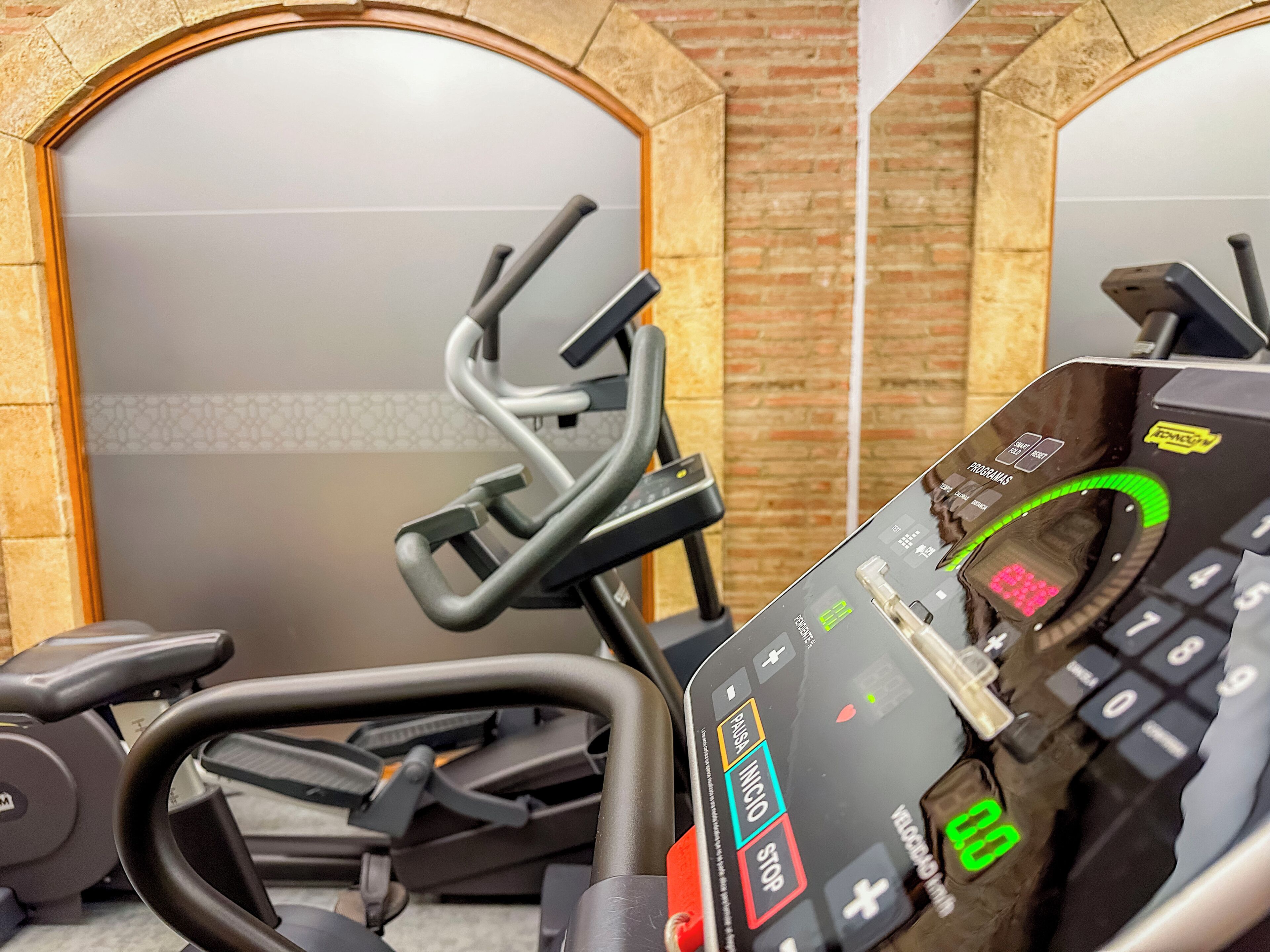 En motionscykel med en digital display och en kontrollpanel, placerad i ett rum med tegelväggar och ett klinkergolv.