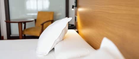 A neatly made bed with white linens and a single white pillow, a wooden headboard, and a small table with a lamp in a hotel room.